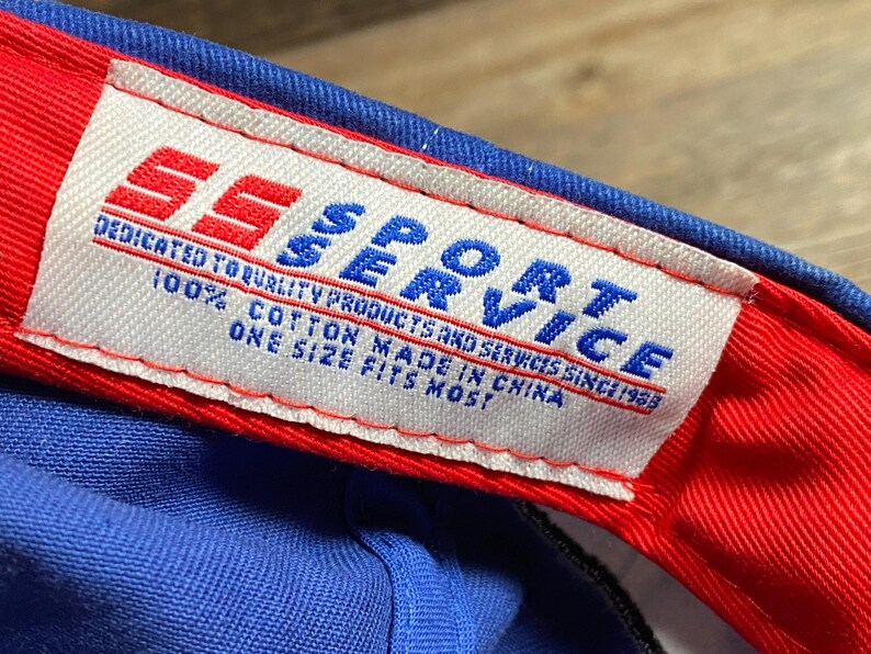 Puede incluir: Primer plano de una gorra de b&eacute;isbol azul con borde rojo. La etiqueta interior dice "SS SPORT SERVICE" con texto que indica que la gorra es 100% algod&oacute;n y talla &uacute;nica.