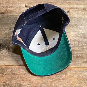 May include: A navy blue baseball cap with a snapback closure, featuring a bright green underside on the brim, white interior panels, dark blue structural stitching, and a small white fabric tag attached to the inner side seam of the crown.