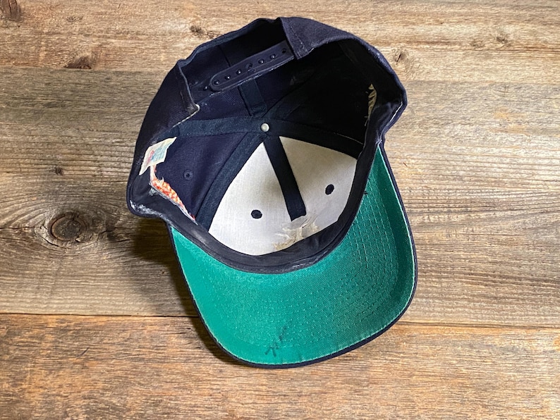 May include: A navy blue baseball cap with a snapback closure, featuring a bright green underside on the brim, white interior panels, dark blue structural stitching, and a small white fabric tag attached to the inner side seam of the crown.