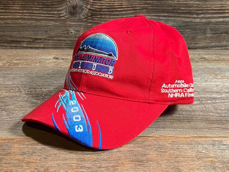 Op de afbeelding: Een felrode honkbalpet met een geborduurd logo van de Top Eliminator Club National Hot Rod Association. De pet heeft blauwe grafische accenten, het jaartal 2003 op de klep en de tekst 39th Automobile Club Southern California NHRA Finals aan de zijkant.