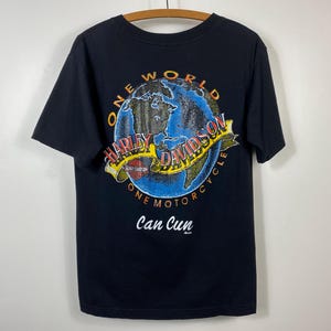 May include: Black t-shirt featuring a colorful graphic of a globe with the words "ONE WORLD HARLEY-DAVIDSON ONE MOTORCYCLE" and "Can Cun". The design includes a yellow banner with the Harley-Davidson logo.