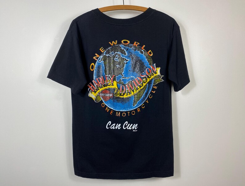 May include: Black t-shirt featuring a colorful graphic of a globe with the words "ONE WORLD HARLEY-DAVIDSON ONE MOTORCYCLE" and "Can Cun". The design includes a yellow banner with the Harley-Davidson logo.