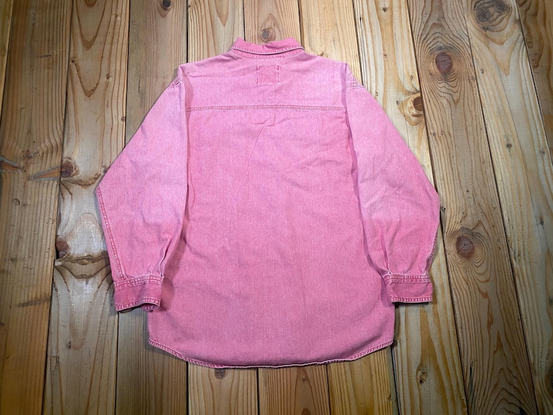 Puede incluir: Una camisa de manga larga con botones en un color rosa s&oacute;lido. La prenda presenta un cuello cl&aacute;sico, un canes&uacute; horizontal en la espalda y pu&ntilde;os abotonados al final de cada manga. La tela tiene una apariencia texturizada con costuras visibles.