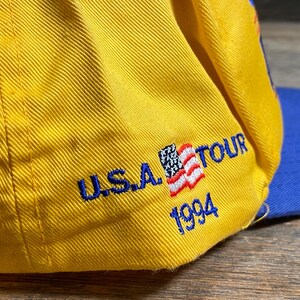 May include: A yellow baseball cap with a blue brim. The cap features the text "U.S.A. TOUR 1994" embroidered in blue, along with a small American flag patch.