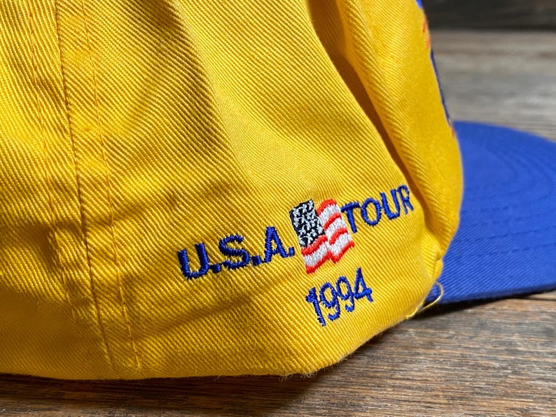 May include: A yellow baseball cap with a blue brim. The cap features the text "U.S.A. TOUR 1994" embroidered in blue, along with a small American flag patch.