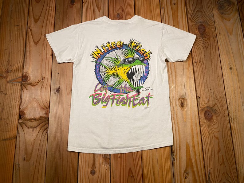 Puede incluir: Camiseta blanca con un gr&aacute;fico colorido de un pez de dibujos animados con gafas de sol y el texto "Nitro Fish". La frase "Let the Big Fish Eat" est&aacute; impresa debajo. La camiseta es de manga corta.