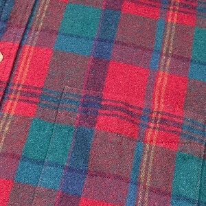 May include: Close-up of a red, green, blue, and brown plaid flannel shirt. The shirt has a button-down front with two visible buttons and a pocket. The plaid pattern is a classic design, with a mix of colors.