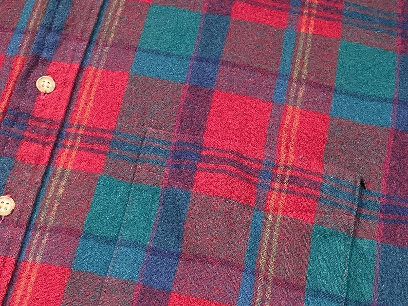May include: Close-up of a red, green, blue, and brown plaid flannel shirt. The shirt has a button-down front with two visible buttons and a pocket. The plaid pattern is a classic design, with a mix of colors.