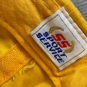 May include: Close-up of a yellow fabric with a sewn seam and a rectangular label. The label has the text "SS SPORT SERVICE" in blue and red, with gold-colored curved lines.