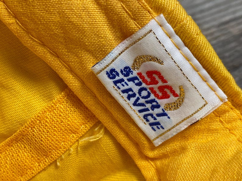 May include: Close-up of a yellow fabric with a sewn seam and a rectangular label. The label has the text "SS SPORT SERVICE" in blue and red, with gold-colored curved lines.