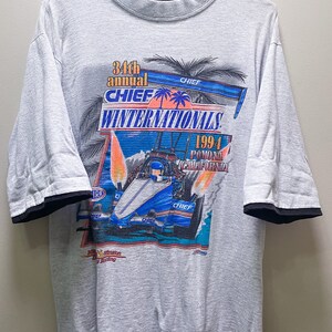 May include: A gray t-shirt with a black mock neck collar and black trim on the sleeves. The front of the shirt features a graphic of a race car with flames and palm trees. The text "34th annual CHIEF WINTERNATIONALS 1994 Pomona California" is printed on the shirt.