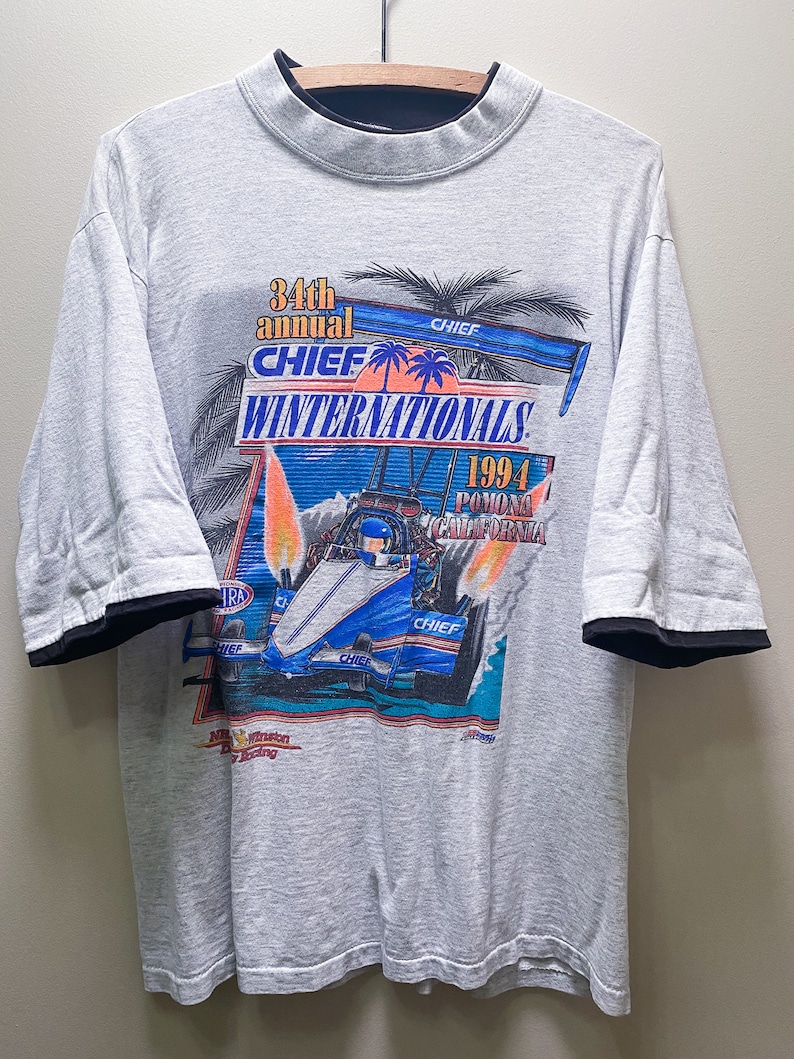 May include: A gray t-shirt with a black mock neck collar and black trim on the sleeves. The front of the shirt features a graphic of a race car with flames and palm trees. The text "34th annual CHIEF WINTERNATIONALS 1994 Pomona California" is printed on the shirt.