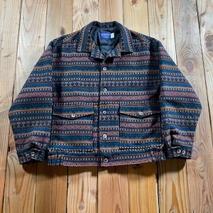 May include: A long-sleeve jacket featuring a multi-coloured geometric pattern in shades of blue, brown, and red. The jacket includes a pointed collar, a front button closure, two large flap pockets on the chest, and an elasticated hem at the waist.