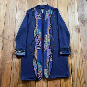May include: A long-sleeved navy blue cardigan featuring a central vertical panel of multicolored floral and geometric patterned fabric. The front opening is framed by dark, wavy trim, and the cuffs are accented with matching patterned fabric.