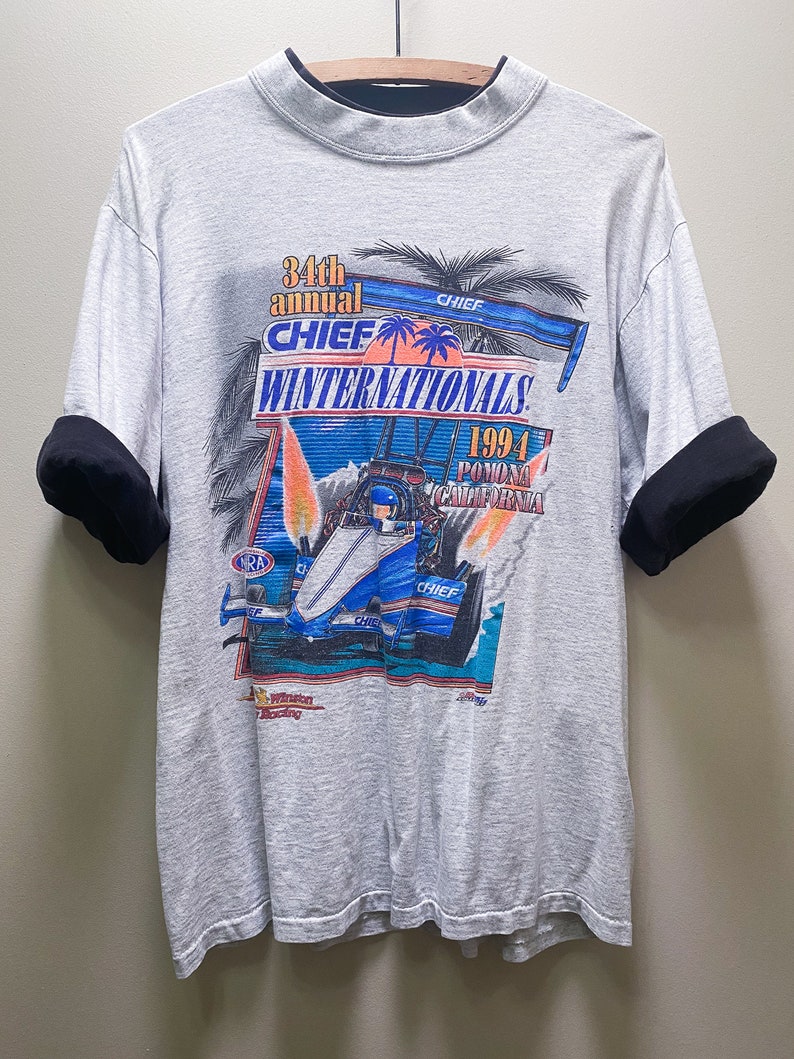 May include: A gray t-shirt with a black and gray collar and black sleeves. The shirt has a graphic print of a race car with the text '34th annual CHIEF WINTERNATIONALS 1994 POMONA CALIFORNIA'.