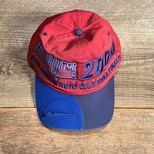 May include: A red baseball cap with a blue brim featuring embroidered text that reads TERMINATOR 2000, 36TH ANNUAL AUTO CLUB NHRA FINALS. The cap has a blue embroidered graphic of a race car on the left side of the brim and a blue button on top.