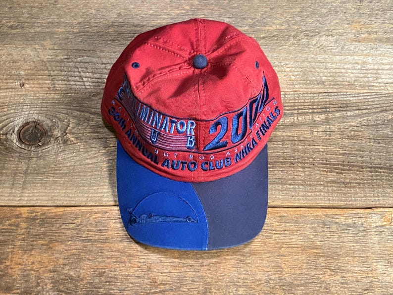 May include: A red baseball cap with a blue brim featuring embroidered text that reads TERMINATOR 2000, 36TH ANNUAL AUTO CLUB NHRA FINALS. The cap has a blue embroidered graphic of a race car on the left side of the brim and a blue button on top.