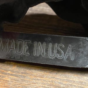 May include: Close-up of a black hat strap with the embossed text "MADE IN USA". The strap is made of a textured, dark material and is attached to a black hat. The background is a wooden surface.