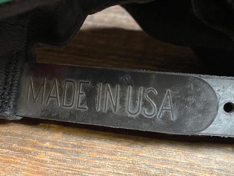 May include: Close-up of a black hat strap with the embossed text "MADE IN USA". The strap is made of a textured, dark material and is attached to a black hat. The background is a wooden surface.