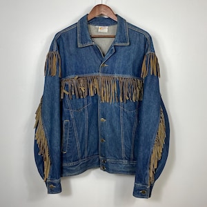 May include: A vintage denim jacket in a medium blue wash, featuring brown leather fringe detailing on the sleeves and chest. It has a classic collar, button-down front, and two chest pockets. The jacket is hung on a wooden hanger.