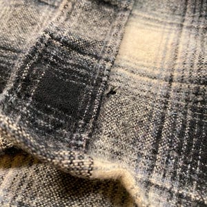 May include: Close-up of a section of plaid fabric in shades of grey, black, and off-white. The fabric has a textured appearance with visible woven threads. The pattern is a classic check design, with the fabric folded over.