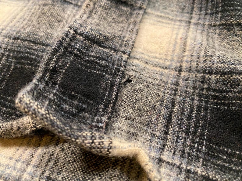 May include: Close-up of a section of plaid fabric in shades of grey, black, and off-white. The fabric has a textured appearance with visible woven threads. The pattern is a classic check design, with the fabric folded over.