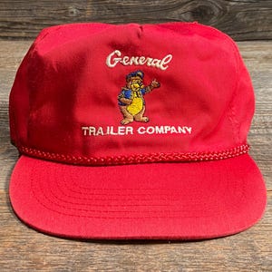 Op de afbeelding: Rode baseballcap met de woorden "General Trailer Company" in het wit geborduurd. De pet heeft een cartoon berenafbeelding. De klep is effen rood en de pet heeft een gevlochten touwdetail rond de basis.