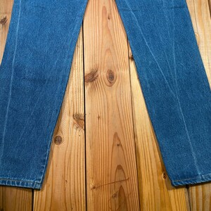 May include: A pair of blue denim jeans, laid flat on a wooden surface. The jeans are a medium wash, with visible seams and a classic straight-leg cut. The fabric shows subtle wear patterns.