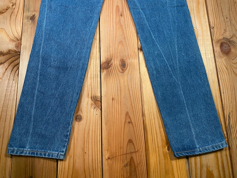 May include: A pair of blue denim jeans, laid flat on a wooden surface. The jeans are a medium wash, with visible seams and a classic straight-leg cut. The fabric shows subtle wear patterns.