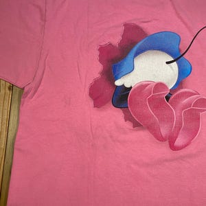 May include: A bright pink t-shirt featuring a graphic print of a cartoon character with a blue hat and pink shoes emerging from a dark purple, jagged, hole-like shape. The character has a white face and a single thin black line extending upward.