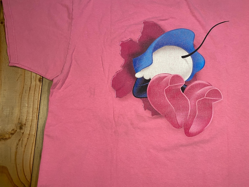 May include: A bright pink t-shirt featuring a graphic print of a cartoon character with a blue hat and pink shoes emerging from a dark purple, jagged, hole-like shape. The character has a white face and a single thin black line extending upward.