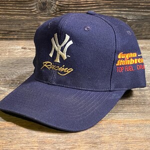 May include: A navy blue baseball cap featuring an embroidered white New York Yankees logo and the word Racing in gold thread on the front. The side displays the embroidered text Gwynn Steinbrenner TOP FUEL DRAGSTER in orange and red lettering.