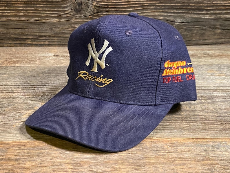 May include: A navy blue baseball cap featuring an embroidered white New York Yankees logo and the word Racing in gold thread on the front. The side displays the embroidered text Gwynn Steinbrenner TOP FUEL DRAGSTER in orange and red lettering.