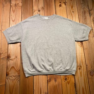 May include: A short-sleeved sweatshirt in a heather gray color featuring a crew neckline with a V-shaped stitch detail at the collar. The garment has ribbed cuffs on the sleeves and a ribbed hemline at the bottom, constructed from a soft knit fabric.