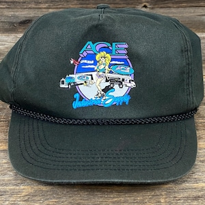 May include: Dark green trucker hat with a black rope detail. The front features a colorful graphic of a woman, trucks, and the text "ACE Industrial Supply". The hat has a curved brim and a mesh back.