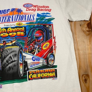 May include: White t-shirt featuring a colorful graphic of a drag racing car with the text "CHIEF WINTERNATIONALS 38th Annual 1998" and "POMONA CALIFORNIA". The design includes palm trees, a driver in a helmet, and sponsor logos.