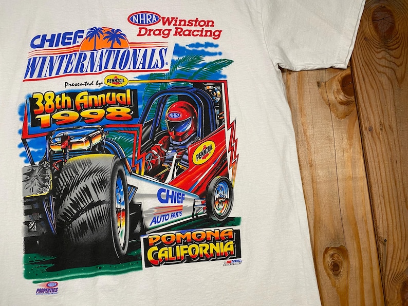 May include: White t-shirt featuring a colorful graphic of a drag racing car with the text "CHIEF WINTERNATIONALS 38th Annual 1998" and "POMONA CALIFORNIA". The design includes palm trees, a driver in a helmet, and sponsor logos.