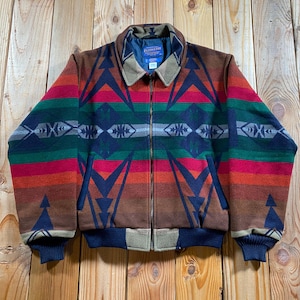 May include: A Pendleton zip-up jacket featuring a multi-colored geometric pattern in shades of brown, red, green, and navy blue. The jacket has a tan collar, a full-length front zipper, two side pockets, and dark navy ribbed cuffs and hem.