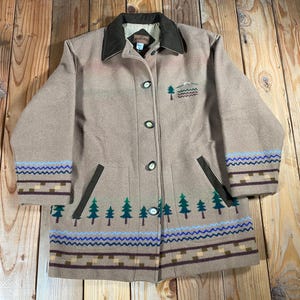 May include: A tan wool-blend jacket featuring a dark brown collar, long sleeves, and a button-front closure. The garment displays embroidered green pine trees, blue wavy lines, and geometric patterns along the hem and sleeve cuffs.