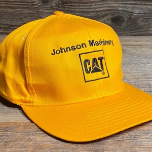 May include: A bright yellow baseball cap with a curved brim. The cap features the text "Johnson Machinery" above a black square with the "CAT" logo.