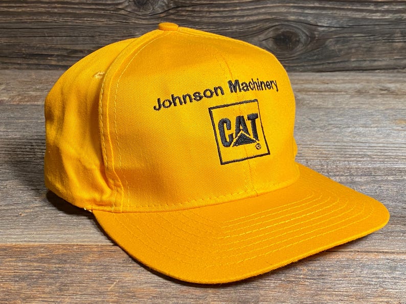 May include: A bright yellow baseball cap with a curved brim. The cap features the text "Johnson Machinery" above a black square with the "CAT" logo.