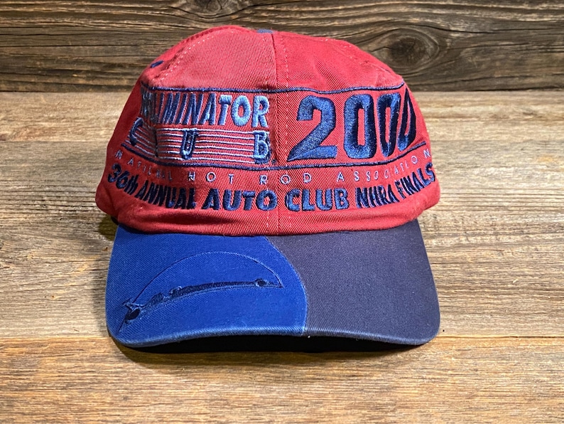 May include: A red baseball cap with a blue brim featuring embroidered text that reads Eliminator Club 2004 and 36th Annual Auto Club NHRA Finals. The cap has a structured crown and a two-toned blue brim with a stitched graphic detail on the left side.