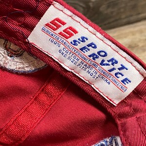 May include: A red baseball cap interior featuring a white fabric tag with the red and blue text SS SPORT. The tag includes the phrases DEDICATED TO QUALITY PRODUCTS AND SERVICES SINCE 1983, 100% COTTON MADE IN CHINA, and ONE SIZE FITS MOST.