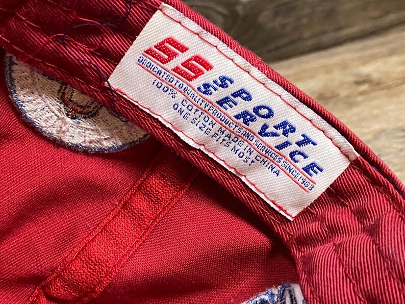 May include: A red baseball cap interior featuring a white fabric tag with the red and blue text SS SPORT. The tag includes the phrases DEDICATED TO QUALITY PRODUCTS AND SERVICES SINCE 1983, 100% COTTON MADE IN CHINA, and ONE SIZE FITS MOST.