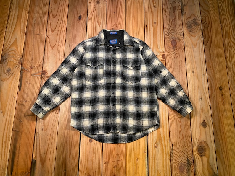 May include: A black and white checked shirt with two chest pockets. The long-sleeved shirt has a classic collar. The fabric looks like a wool blend. The shirt is laid flat on a wooden surface.