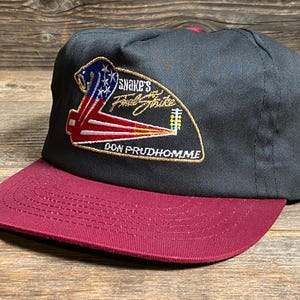 May include: A black baseball cap with a maroon peak. The cap has an embroidered patch with the text "Snake's Final Strike" and "Don Prudhomme" in gold, plus a patriotic design in red, white, and blue.