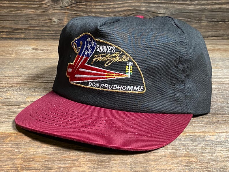 May include: A black baseball cap with a maroon peak. The cap has an embroidered patch with the text "Snake's Final Strike" and "Don Prudhomme" in gold, plus a patriotic design in red, white, and blue.