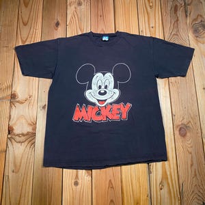 Puede incluir: Una camiseta negra de manga corta con un gr&aacute;fico blanco del rostro de Mickey Mouse con textura desgastada. Debajo del rostro del personaje, la palabra MICKEY est&aacute; impresa en letras may&uacute;sculas rojas, gruesas y con un borde negro.
