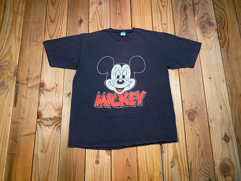 Puede incluir: Una camiseta negra de manga corta con un gr&aacute;fico blanco del rostro de Mickey Mouse con textura desgastada. Debajo del rostro del personaje, la palabra MICKEY est&aacute; impresa en letras may&uacute;sculas rojas, gruesas y con un borde negro.