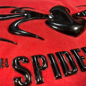 Op de afbeelding: Close-up van een rode stof met een glanzend zwart ontwerp. Het ontwerp bevat het woord "SPIDER" in vette letters en een gestileerde hartvorm. De stof heeft een getextureerd uiterlijk.